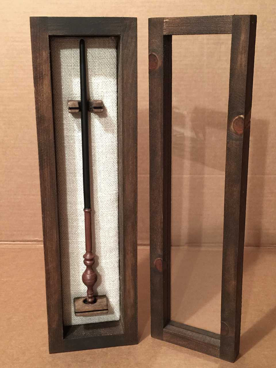 A wooden wand collector's display case with a glass lid and burlap backing with Harry Potter wand, displayed against a tan background.
