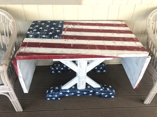 An outdoor rectangular accent table with a weathered American flag design on the tabletop and a white base with blue stars detail. The table is set on a wooden deck with outdoor furniture around it.
