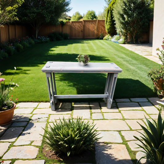 A contemporary bistro table made of solid thermally modifed wood, placed on a paved outdoor area with a plant on top, surrounded by greenery and a wooden fence in the background.