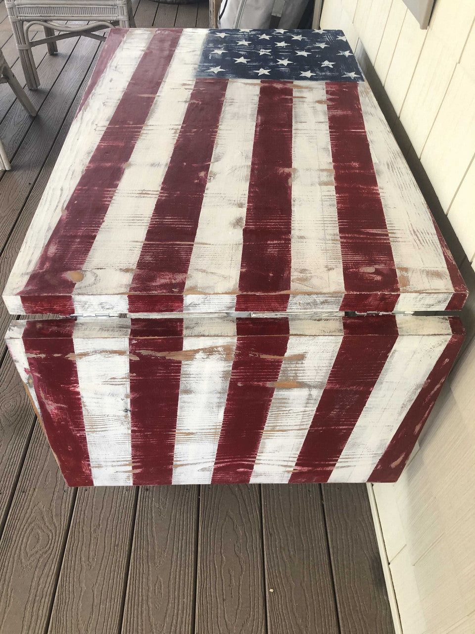 Close-up of outdoor solid wood accent table with a weathered American flag design on the tabletop. The table is set on a wooden deck with outdoor furniture around it.