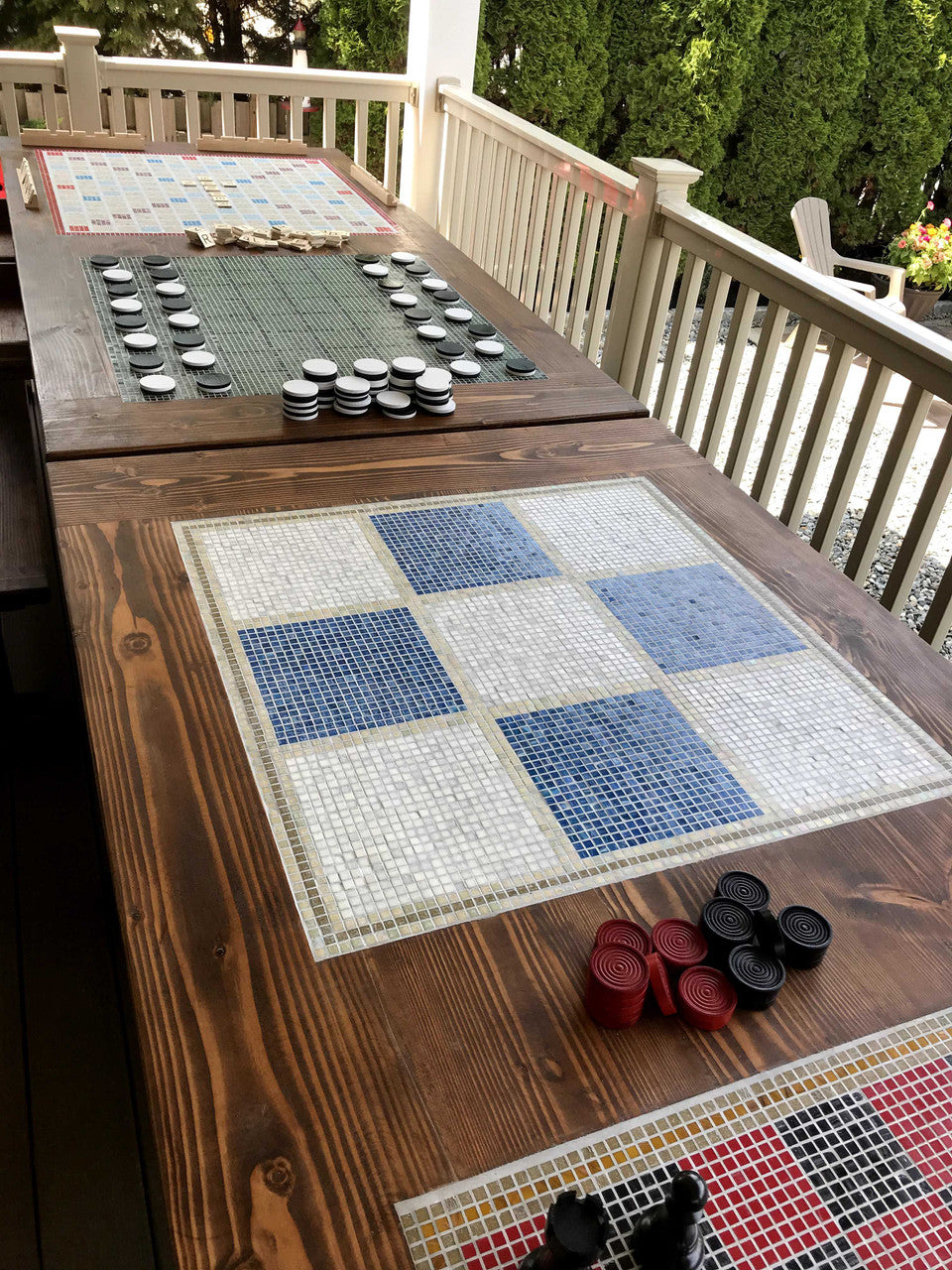 Overhead view of outdoor dining table with inlaid tile gameboards - checkers, tic-tac-toe, othello on elevated deck with green trees in background.