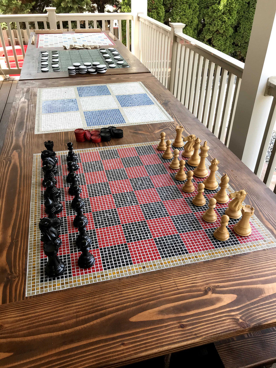 Overhead view of FSC-certified solid wood dining set with inlaid chess board and tik tak toe board in the center, placed on an outdoor deck with green trees in the background.