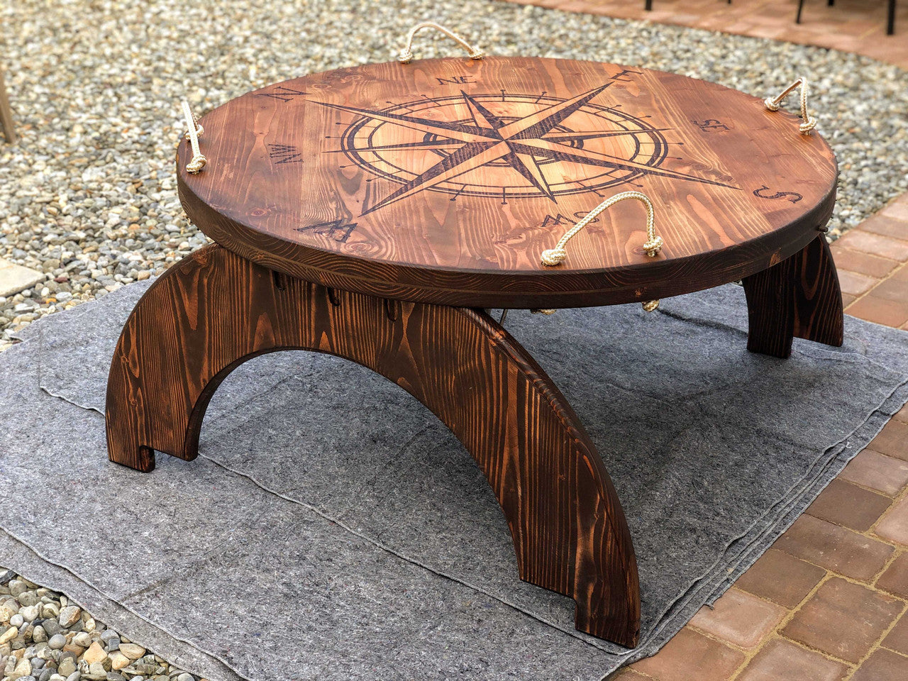 A solid wood fire pit cover with a nautical theme, featuring an oversized compass design, converted into a table when not in used, placey on stone and gravel.