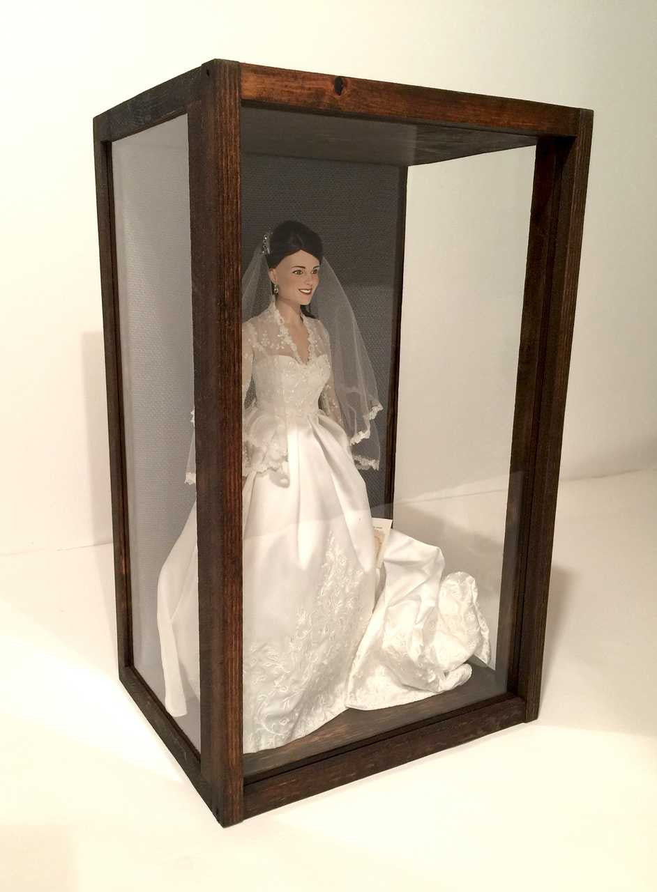 A side view of solid wood 3-sided glass display with a grey burlap backing featuring Princess Kate collectible doll (not included) against a white background.