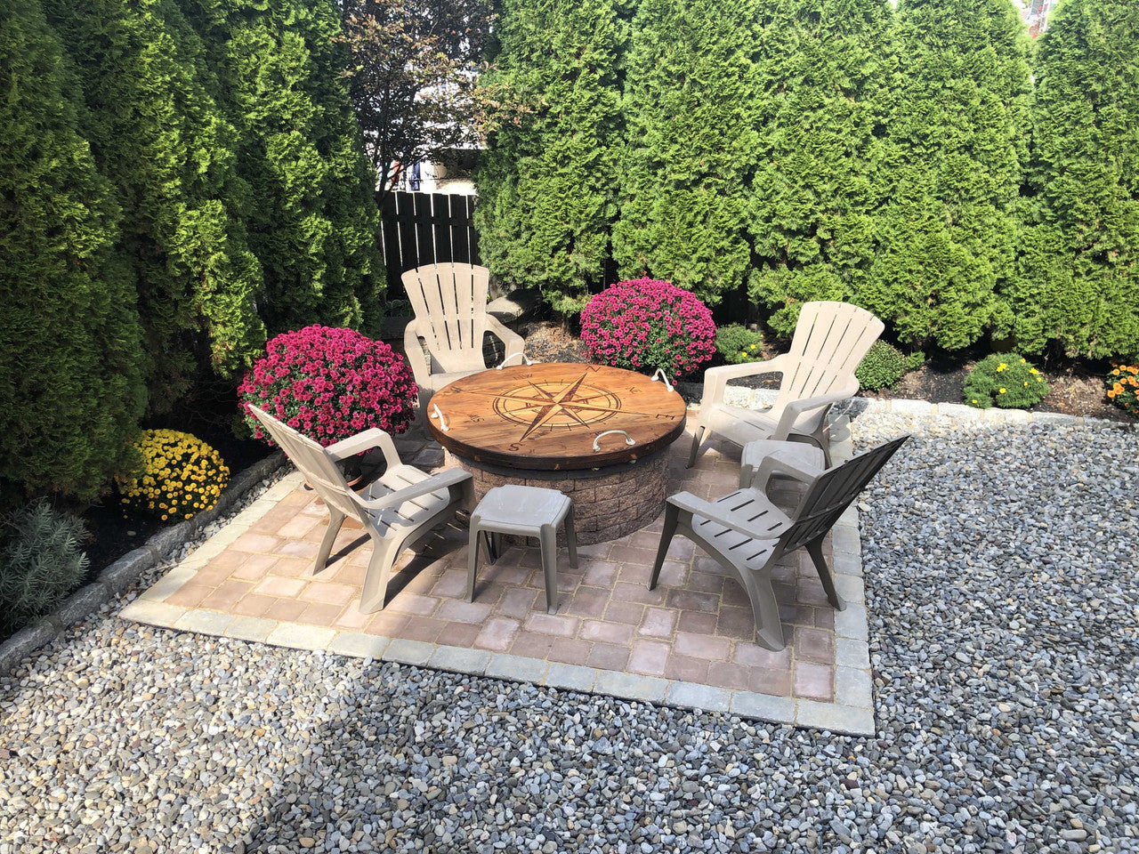A solid wood fire pit cover with a nautical theme,on top of a fire pit featuring an oversized compass design and mooring rope handles placed on a stone platform surrounded by rocks with potted plants and greenery in the background.
