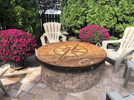 A solid wood fire pit cover with a nautical theme, featuring an oversized compass design, placed on a stone deck surrounded by chairs, big plants and greenery in the background.