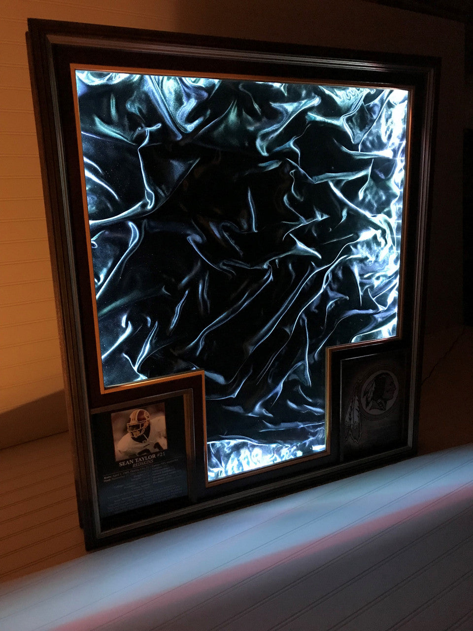 Illuminated view of sports jersey display case featuring a lushb black satin backing, a biopic and a burned team logo,with a magnetic glass lid on a white background.