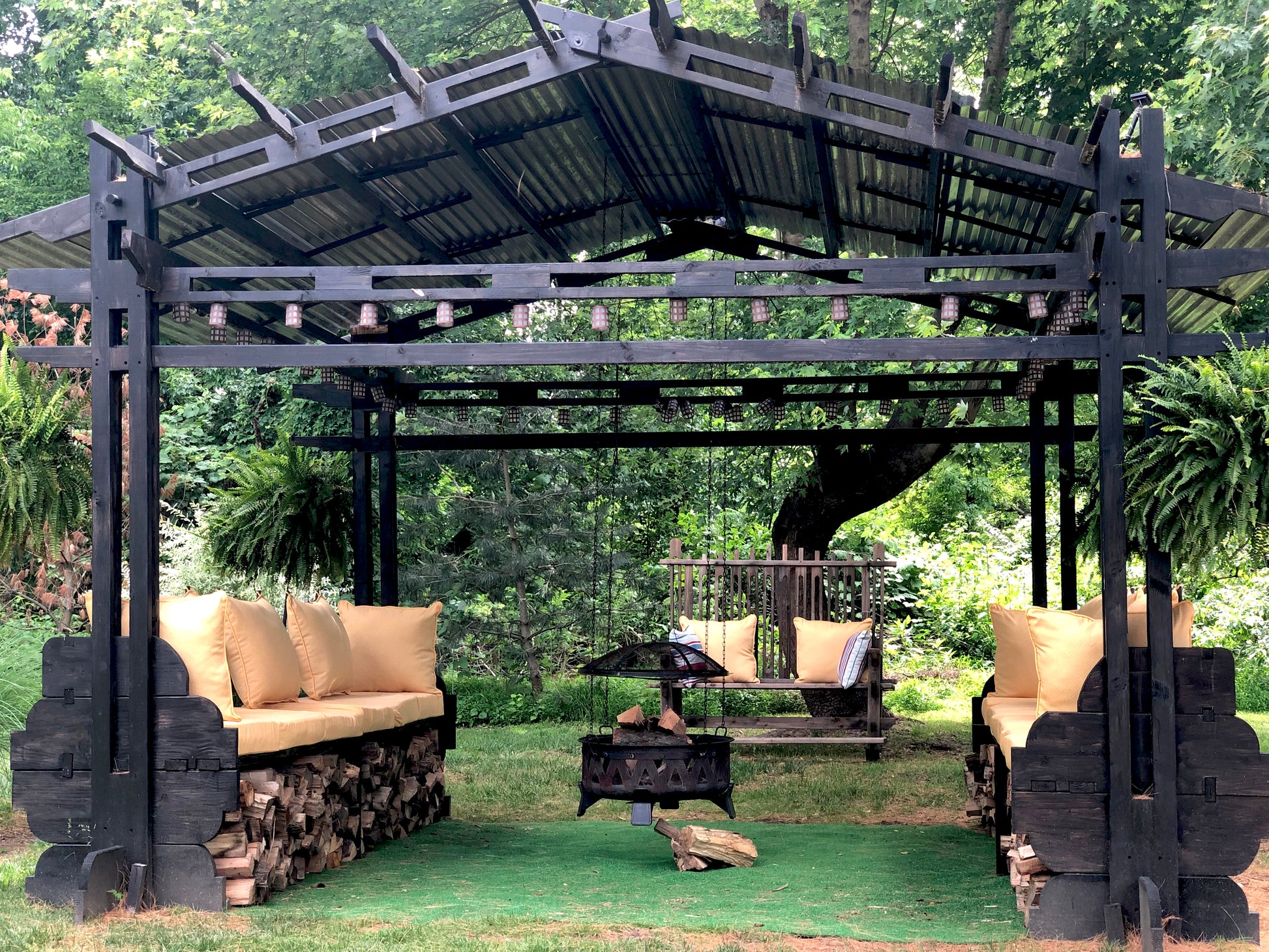 A black solid wood contemporary gazebo with a galvanized steel roof, featuring two oversized lounge-like couches and a hanging fire pit with an oversized outdoor rocking chair in background. Hardware-free, tool-free assembly.