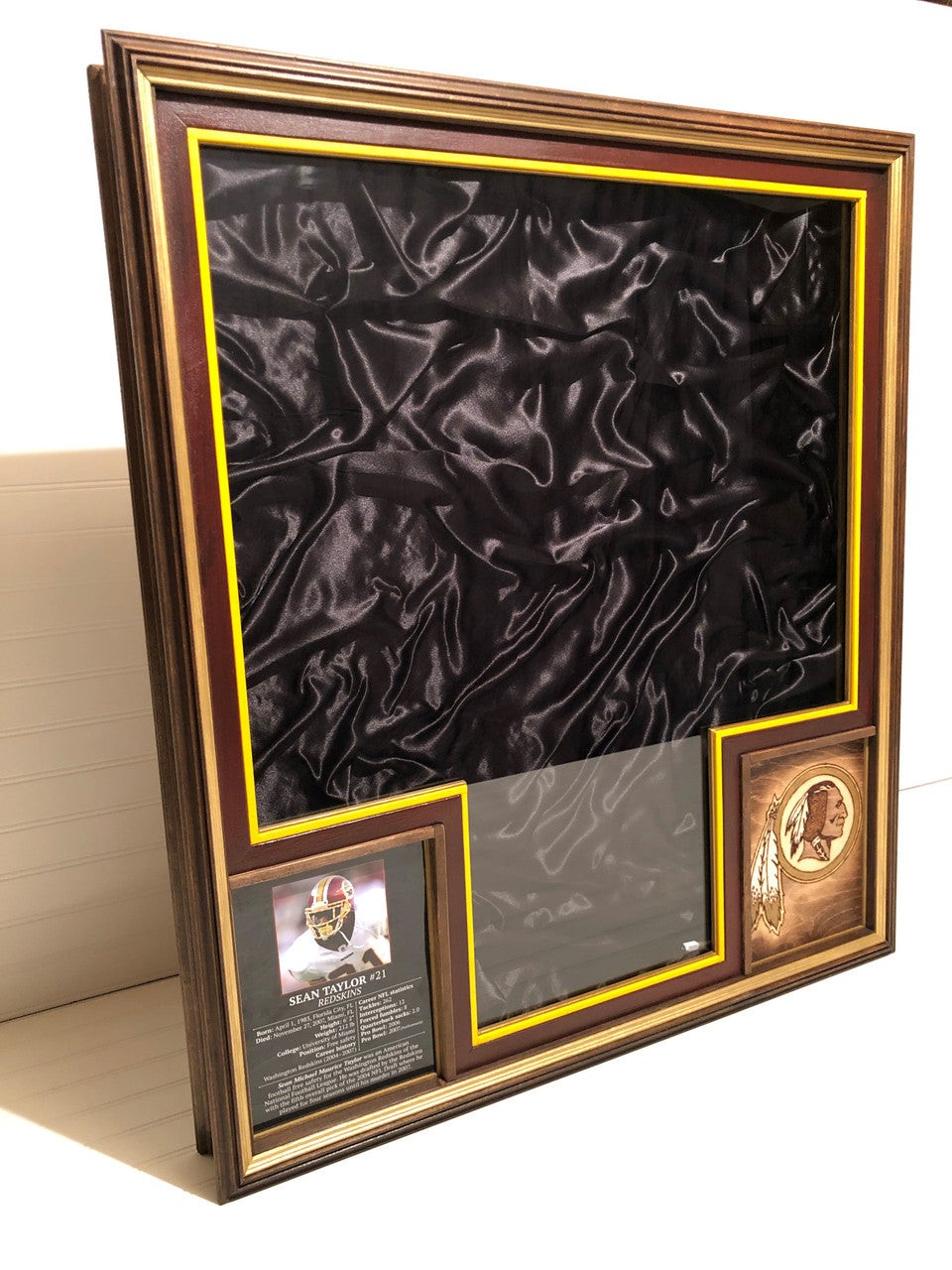 A sports jersey display case featuring a lush satin backing, a biopic and a decorative element with a burned and enameled team logo,with a magnetic glass lid on a white background.