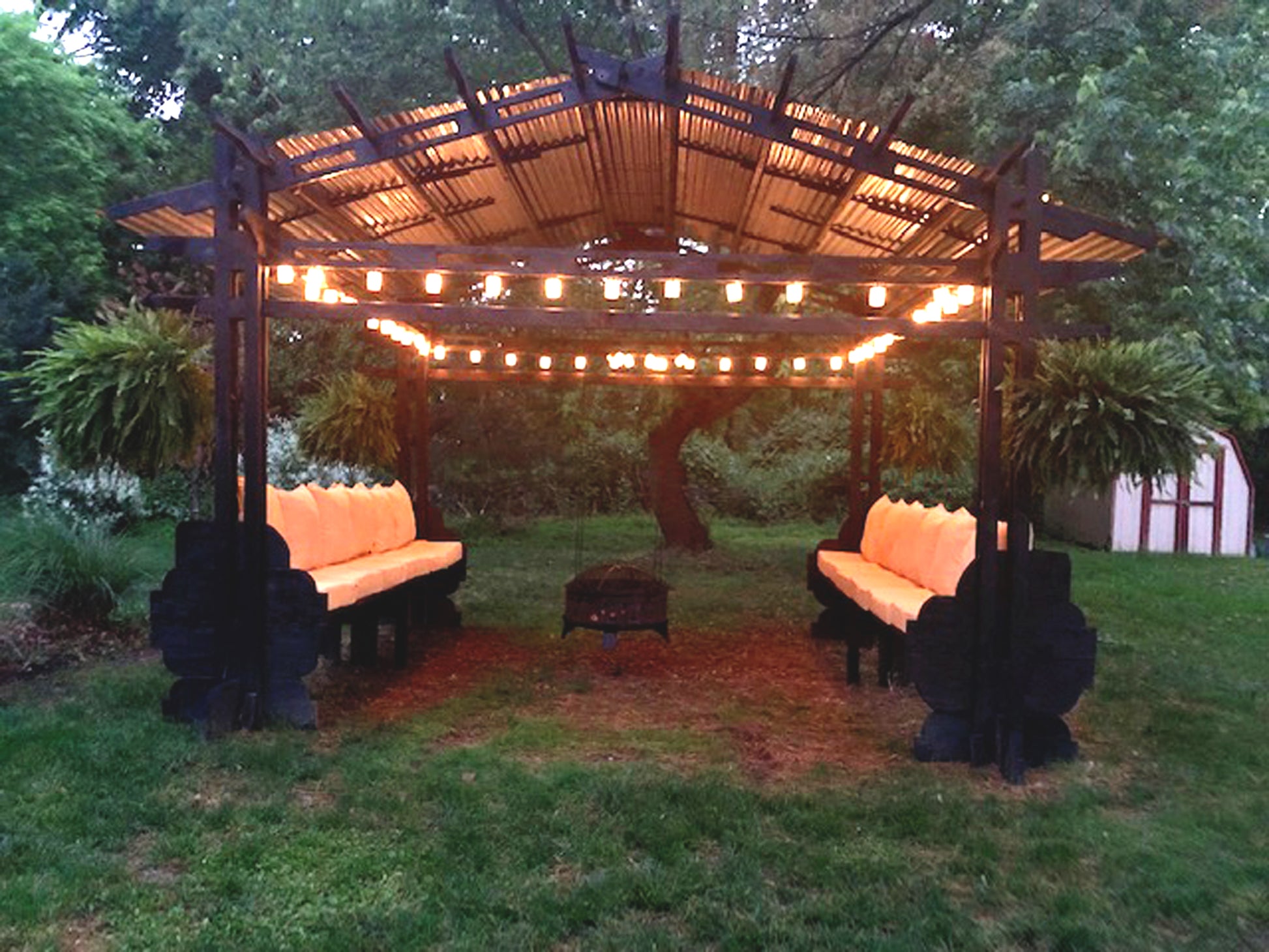 A solid fsc-certified wood gazebo with a galvanized steel roof, featuring two oversized lounge-like couches, hanging fire pit with string lights.