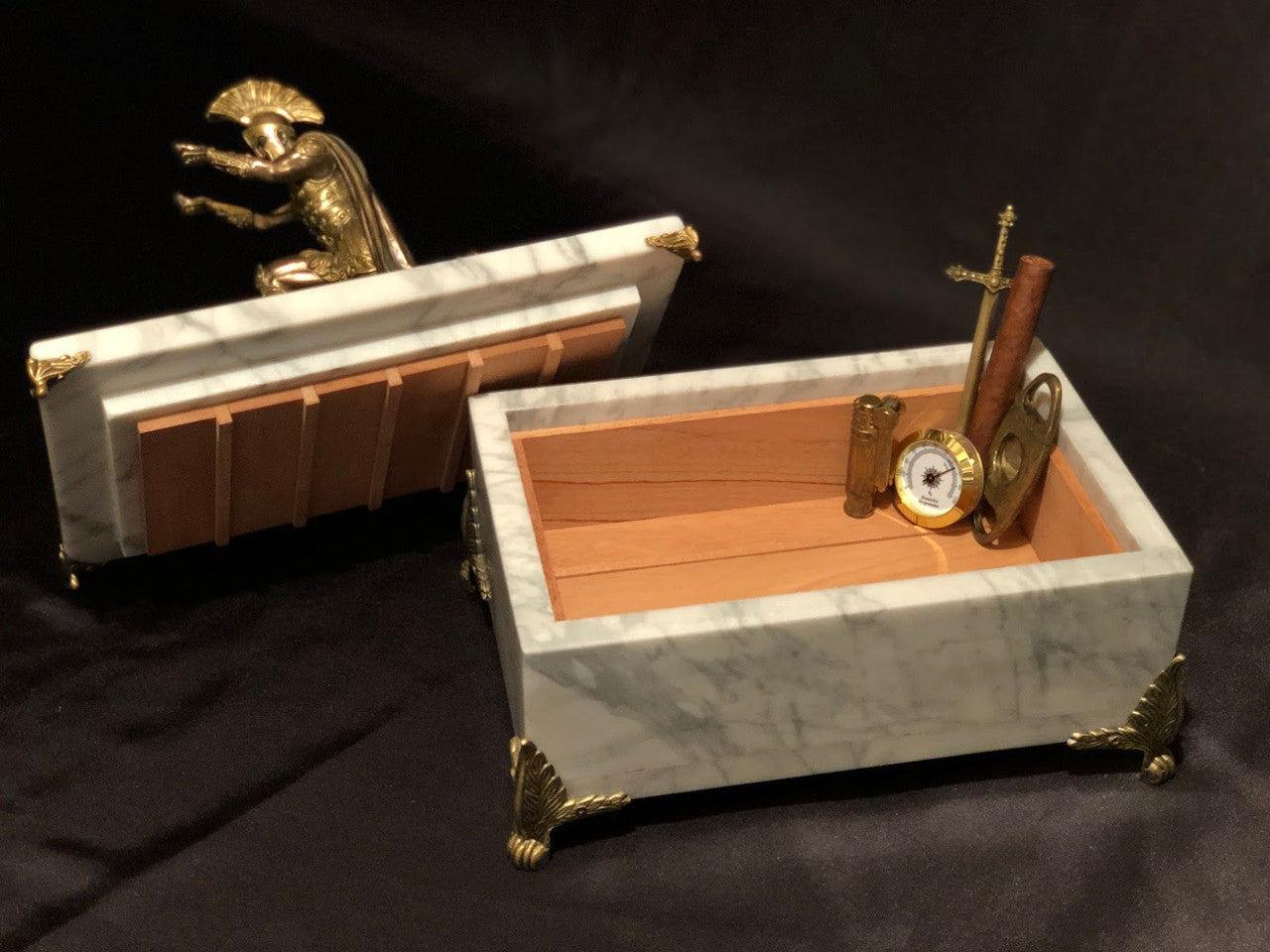 A White Carrara Marble Humidor featuring an antiqued Spartan statue lid handle afixed to marble plinth, with cedar lined interior, cigar, hygrometer, cigar cutter and lighter on a black satin background.